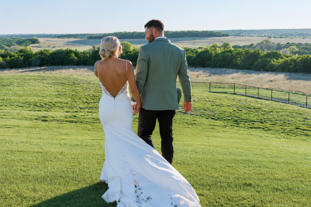 Newlywed couple walking hand in hand across the grounds of Diamond A Ranch with sweeping Texas countryside views and open skies in the background.